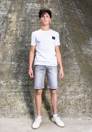 Adolescent garçon aux cheveux foncés se tient contre un mur en béton texturé, portant un t-shirt blanc, un short en jean clair et des baskets blanches avec des accents bleus.