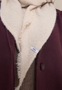 Krémový šál s třásněmi a štítkem "CECIL" schovaným uvnitř bordó kabátu přes texturovaný hnědý svetr.