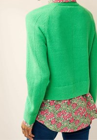 Maglione verde lavorato a maglia con orlo a costine, indossato sopra una camicia a stampa floreale con fiori rosa su uno sfondo verde.