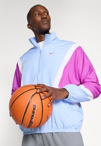 Man in lichtblauw en paars jack die een oranje basketbal vasthoudt en opzij kijkt tegen een effen achtergrond.