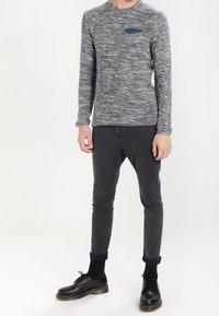 Haut en tricot gris texturé à manches longues avec un détail de poche bleue, associé à un pantalon gris slim-fit et des chaussures noires, avec des poignets retroussés.