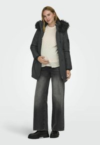 Parka de maternité en gris foncé avec une capuche en fausse fourrure, associée à un pull léger texturé et un jeans large noir. Des bottines noires à la cheville complètent le look.