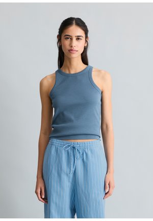Kobieta stoi, ubrana w niebieski prążkowany top bez rękawów i luźne jasnoniebieskie spodnie w paski z wiązaniem na tle jednolitego szarego tła.