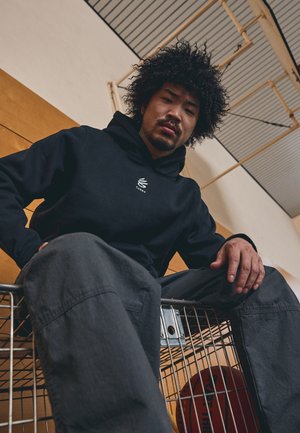 Sort hoodie med et hvidt logo på brystet, parret med mørkegrå bukser. Model siddende i en metal indkøbsvogn med en basketball indeni.