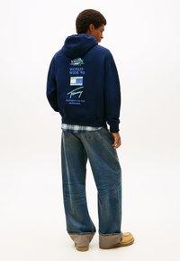 Μπλε φούτερ με κουκούλα σε ναυτικό μπλε χρώμα, με λευκά και πράσινα γραφικά λογότυπα στην πλάτη, συνδυασμένο με χαλαρά τζιν παντελόνια και καφέ μποτάκια.