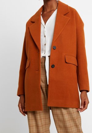 Person iført en rust-orange uldfrakke over en hvid knappet bluse, der er bundet i taljen, og beige bukser med tern.