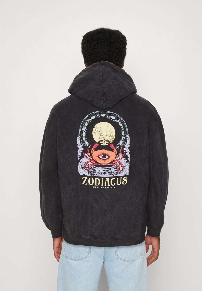 Personne portant un sweat à capuche noir avec une illustration colorée de crabe présentant un œil unique, une pleine lune et le texte "ZODIACUS" dans le dos.