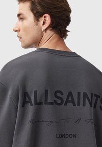 Grå sweatshirt med rund hals, prydet med sort trykt tekst "ALLSAINTS" og "Message To A Friend" på ryggen. Blød tekstur.