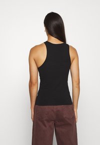 Mujer con cabello oscuro y largo, usando pendientes de aro dorados, una blusa sin mangas de corte deportivo negra y pantalones de tiro alto de pierna ancha de color marrón oscuro.