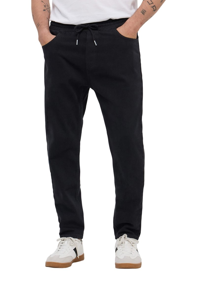 Homme portant un pantalon noir à cordon et des baskets blanches, les mains dans les poches, tatouages visibles sur les avant-bras, debout sur un fond blanc.