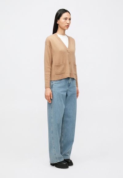 Καρντιγκάν χρώματος ταμπά με V-λαιμόκοψη, κουμπιά στο μπροστινό μέρος και δύο τσέπες, σε συνδυασμό με ανοιχτό μπλε oversized τζιν και μαύρα chunky μποτάκια.