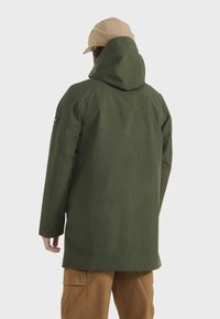 Grüne wasserdichte Parkajacke mit Kapuze, leichtes Material, gerader Schnitt und minimalistisches Design; kombiniert mit sandfarbenen Cargo-Hosen.