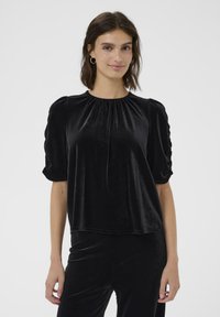 Top in velluto nero con scollatura arricciata, maniche a sbuffo corte e vestibilità facile. Texture liscia con un leggero luccichio. Senza fantasie né accessori.