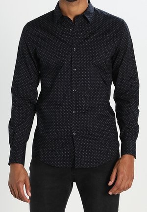 Man draagt een zwart overhemd met knopen en lange mouwen, bedrukt met kleine witte polkadots, gecombineerd met een zwarte broek.