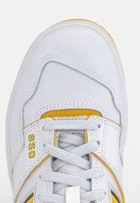 Scarpa sportiva bianca con un upper in pelle testurizzata, lacci grigi, dettagli gialli e un'etichetta "650" visibile vicino alla punta.