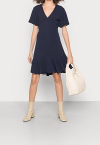 Námořnické v-čkový šaty s krátkými volánkovými rukávy, rozevlátým spodním okrajem a siluetou do A. V kombinaci s béžovými vysokými botami na podpatku a měkkou taškou.
