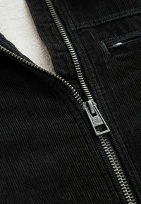 Zwarte corduroy jas met een zilveren rits, voorzien van een zachte witte binnenvoering en een verticale ribbelstructuur op de buitenstof.