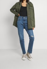 Chaqueta oversized verde oliva con múltiples bolsillos, camiseta negra sin mangas, jeans rectos azules claros con una rodilla rota y zapatillas con estampado de animal.