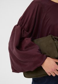 Burgunderbluse med voluminøse, samlede ærmer i et tekstureret stof. Olivengrøn suede clutch holdt i armen, med en blød overflade.