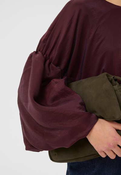 Blouse bourgogne avec des manches volumineuses et froncées en tissu texturé. Pochette en suède vert olive tenue sous le bras, présente une finition douce.