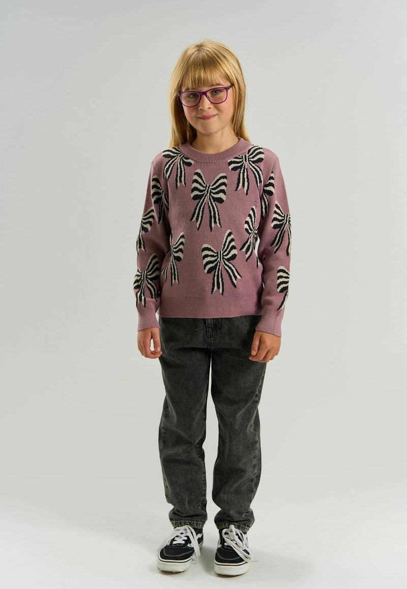 Jeune fille portant des lunettes violettes, un pull violet avec des nœuds noirs et blancs, un jean foncé et des baskets noir et blanc, debout devant un fond uni.