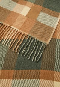 Coperta a scacchi in morbida lana, con quadrati arancioni, verdi, crema e marroni. Include frange texturizzate e un'etichetta in pelle.