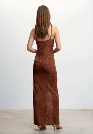 Woman with long brown hair wearing a floor-length, sheer brown floral dress and beige heeled sandals, standing with hands on hips, back facing camera.