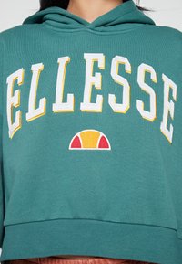 Cropped hoodie in teal realizzato in tessuto morbido, presenta un grande testo bianco "ELLESSE" contornato in giallo e un logo dettagliato in rosso, giallo e bianco.