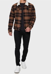 Veste à carreaux marron et noire avec un col en sherpa blanc, des boutons sur le devant et deux poches poitrine ; associée à un pantalon noir et des baskets blanches.