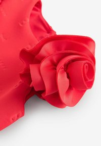 Červený odev s textúrovaným kvetinovým akcentom na pleci, s vrstvenou tkaninou vytvárajúcou 3D efekt. Hladký a jemný materiál.