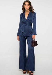 Costume bleu marine à motif floral avec un design enveloppant, pantalons à jambes larges et taille ceinturée. Associé à un petit sac à main bordeaux foncé.