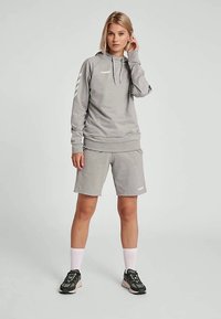Junge Frau steht, trägt einen grauen Kapuzenpullover, passende Shorts, weiße Socken und schwarze Sneakers vor einem schlichten hellen Hintergrund.