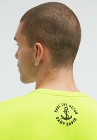 Neon-gelbes T-Shirt mit kurzen Ärmeln und Rundhalsausschnitt, mit schwarzem Anker-Design und dem Text "RACE THE OCEAN CAMP DAVID" auf der Rückseite.