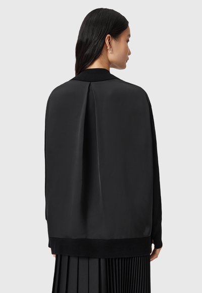 Femme avec des cheveux noirs lisses portant une veste en satin noir ample et une jupe plissée, vue de dos contre un fond gris uni.