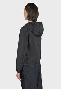 Giacca leggera nera con cappuccio, realizzata in materiale liscio e resistente all'acqua. Presenta polsini elastici e un logo sulla manica sinistra.