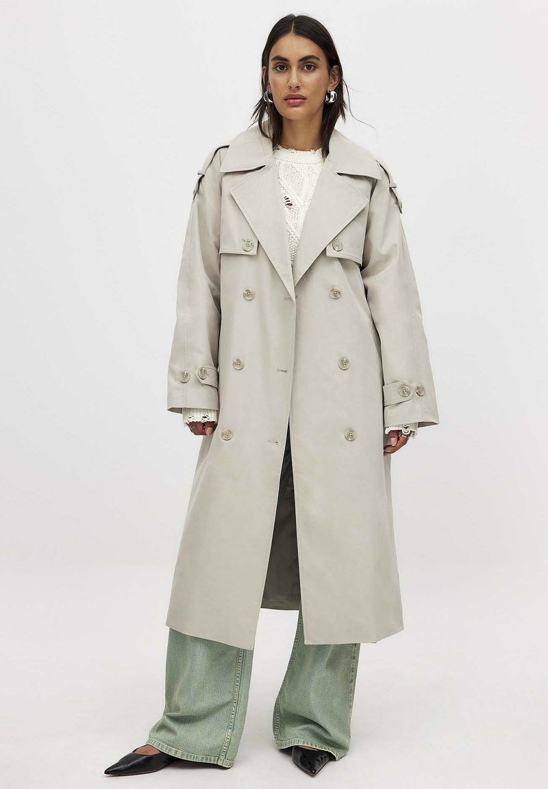 Trench coat doble faz en color beige claro con solapas anchas, detalles de botones y un corte relajado, combinado con un top de punto y jeans verde de pierna ancha.