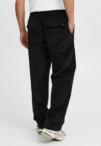 Black corduroy pants with an elastic waistband, subtle texture, and two back pockets, paired with white and gray sneakers.