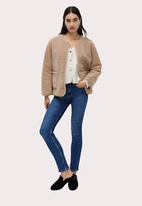 Ljusbeige fleecejacka med rundad krage, framfickor och guldfärgade knappar, matchad med en vit topp och blå skinny jeans. Svarta tofflor.