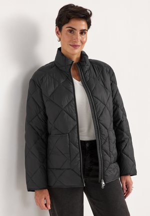 Vrouw die een zwart doorgestikt jack met voorzakken draagt, over een wit shirt en donkere broek, staand tegen een witte muur.