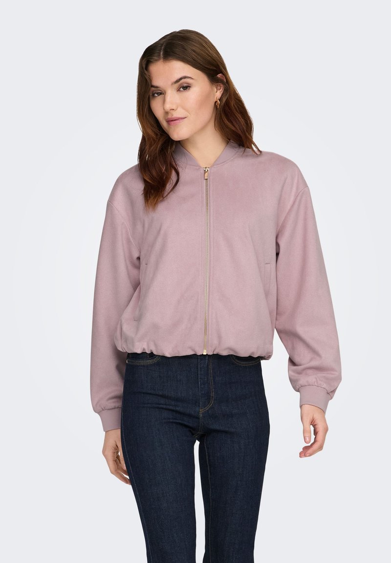 Femme portant une veste bomber zippée violet clair et un jean bleu foncé, debout devant un fond uni clair.