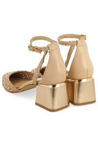 Sandales à talons beiges avec des brides tressées, une boucle à la cheville et des talons en métal. Texture en cuir lisse avec une finition brillante.