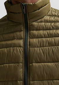 Pružená olivově zelená vesta s vysokým límcem, s černým zipem a prošívanými horizontálními panely pro zvýšení textury.