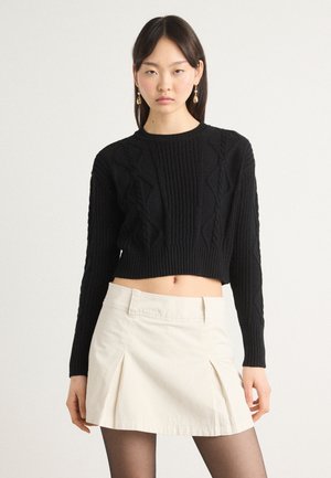 Black knitted crop sweater with ribbed texture and cable patterns, paired with a light beige pleated mini skirt. Model wearing tights.