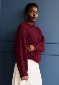 Bordowy sweter z dzianiny warkoczowej o teksturowanym wykończeniu, zaokrąglonym dekolcie i ściągaczach przy mankietach, noszony na kremowej spódnicy na tle niebieskiego tła.