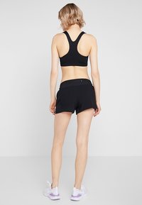 Sujeción deportiva negra con diseño de tirantes en la espalda, combinada con pantalones cortos deportivos negros con una cintura ajustada, mostrada desde la parte de atrás. Zapatillas blancas.