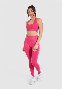 Różowy sportowy biustonosz treningowy z dopasowanymi legginsami, oba z napisem "SMIL". Gładka tekstura, dopasowany krój i elastyczny materiał. Białe tenisówki.