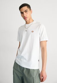 White cotton t-shirt with short sleeves, featuring a small multicolored logo on the left chest. Paired with green cargo pants.