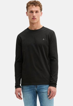 Uomo con capelli ricci che indossa una camicia nera a maniche lunghe e jeans blu, in piedi su uno sfondo chiaro e uniforme.