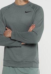 Szara bluza Nike z długim rękawem, okrągłym dekoltem i prążkowanymi mankietami. Posiada czarne logo na górnej lewej części klatki piersiowej. Miękka w dotyku.