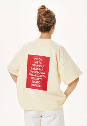 Person mit Dutt trägt ein hellfarbenes, übergroßes T-Shirt mit einem roten Quadrat auf dem Rücken, das italienische Essensnamen auflistet.